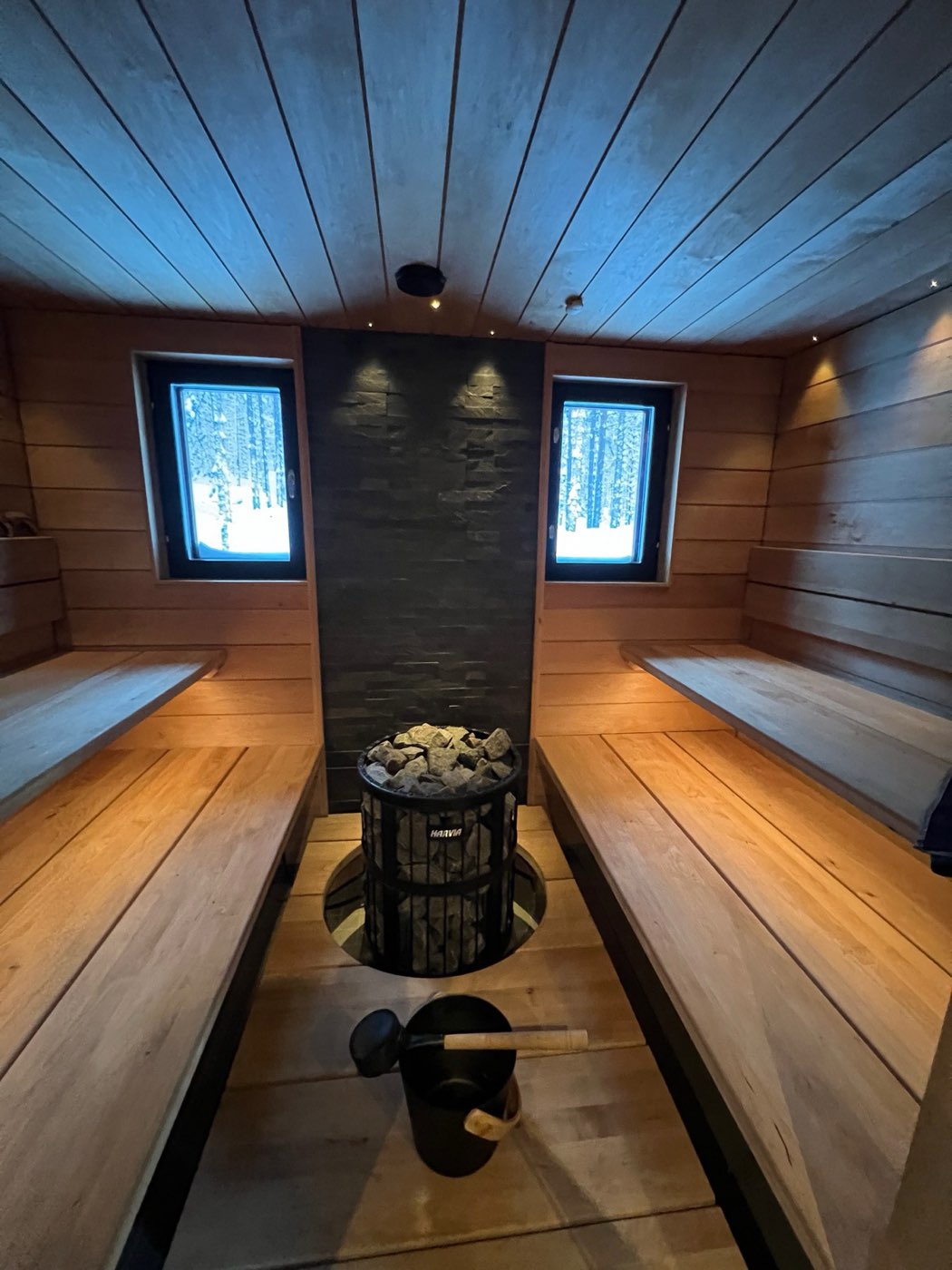 Indoor sauna with wide wooden benches, stone wall, round heater and two windows onto the forest